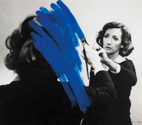 A woman painting thick blue brushstrokes across another person's face, obscuring it